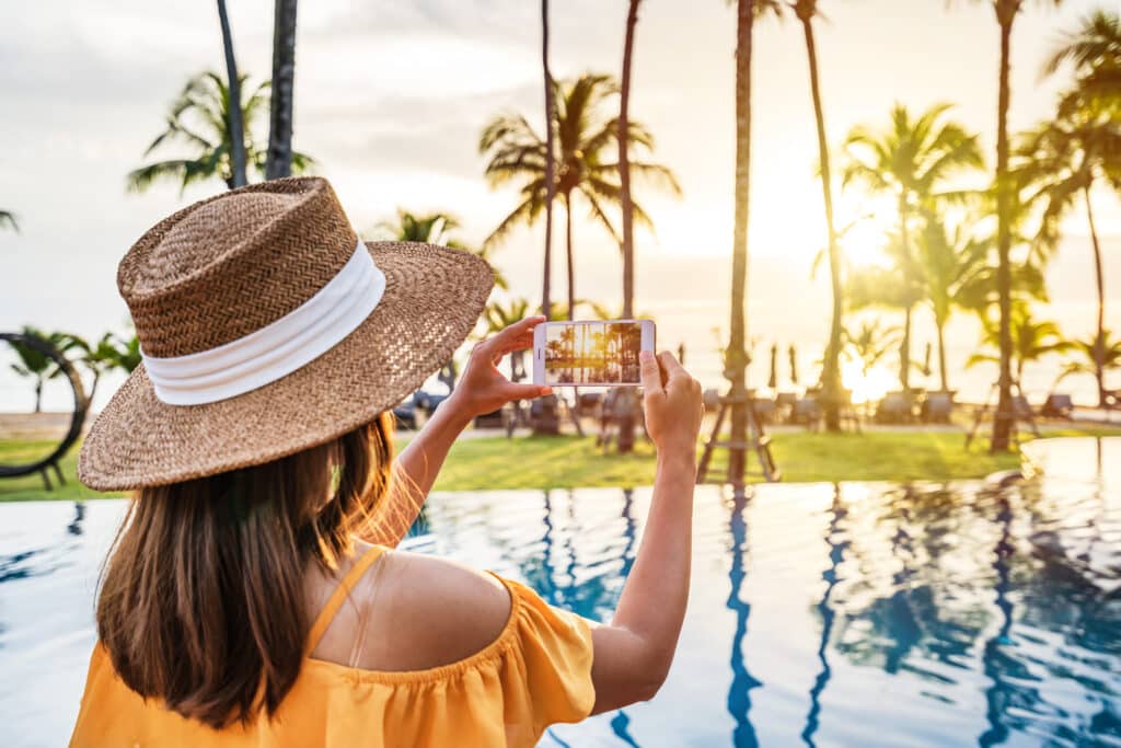 Woman taking a photo of a tropical resort sunset by the pool, capturing vacation moments for social media content